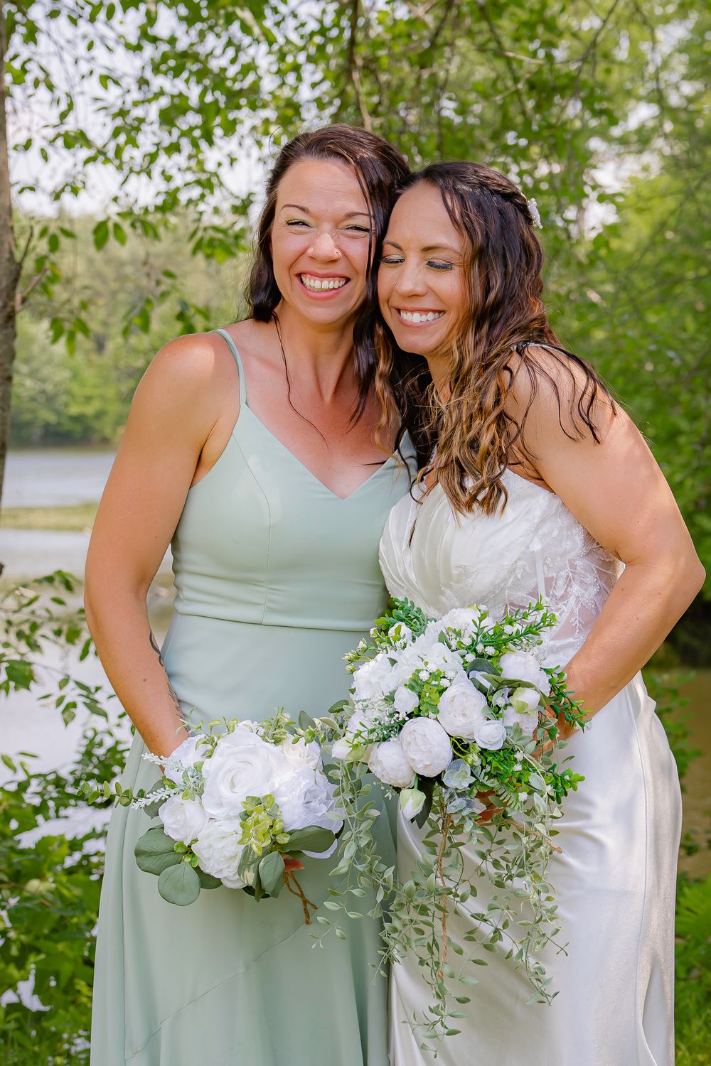 Two people pose and smile together in sage green and ivory dresses while holding white floral bouquets outdoors.