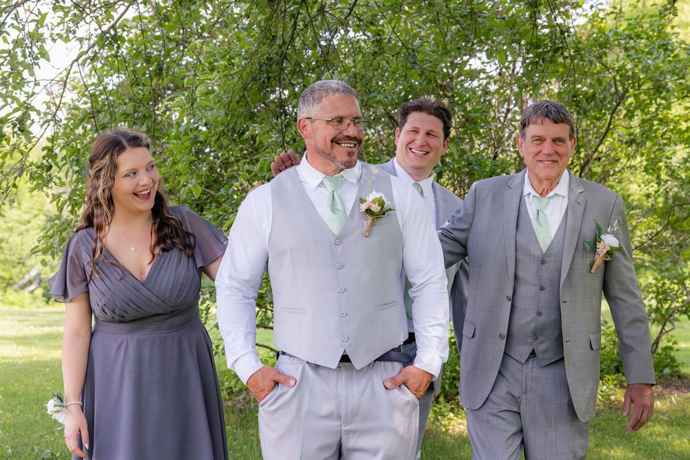 Wedding party walks together while laughing and smiling on grassy path under green trees.