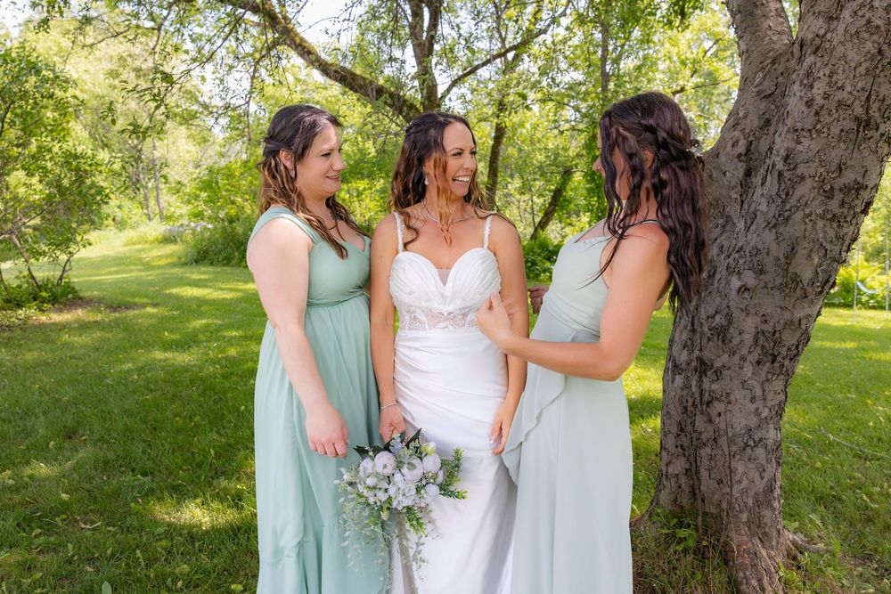 A group wearing sage green and white dresses stand together laughing joyfully under trees at an outdoor celebration.