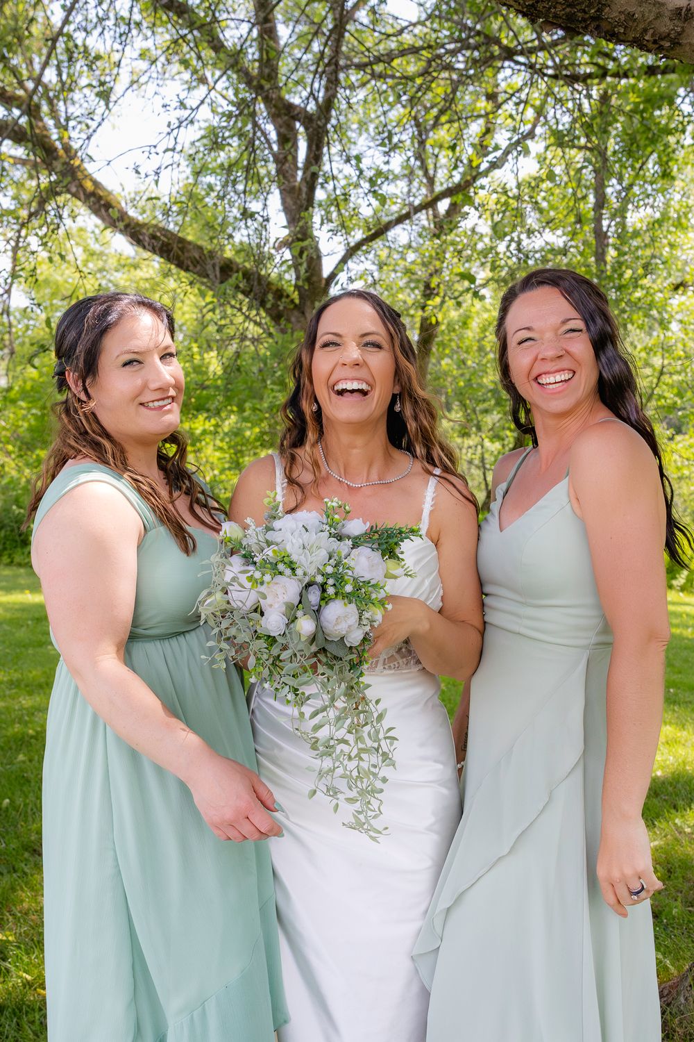 A group wearing sage green bridesmaid dresses and white wedding gown pose together laughing in a sunny orchard.