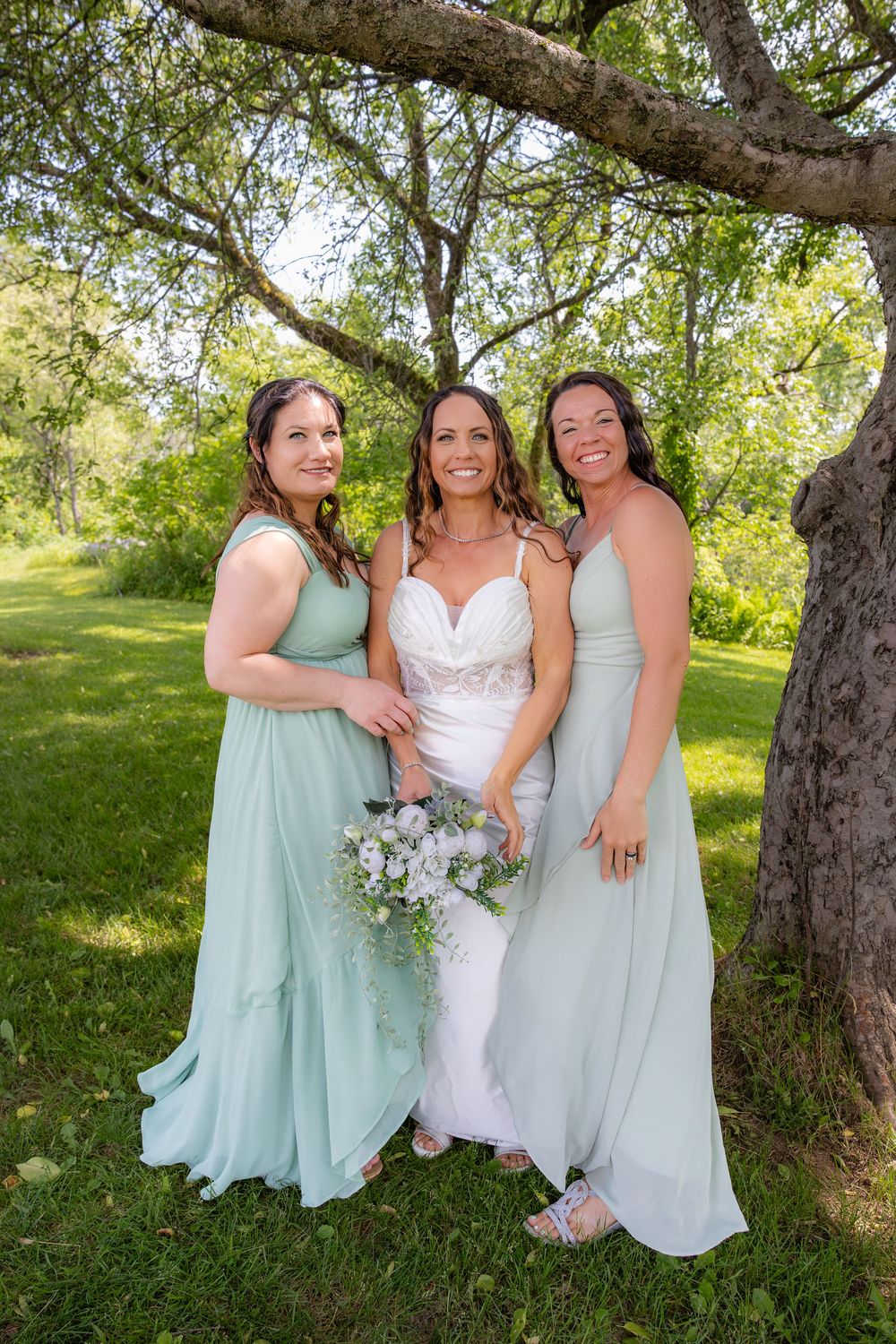 Three people in mint green and white dresses pose together under a tree in a sunny outdoor setting.