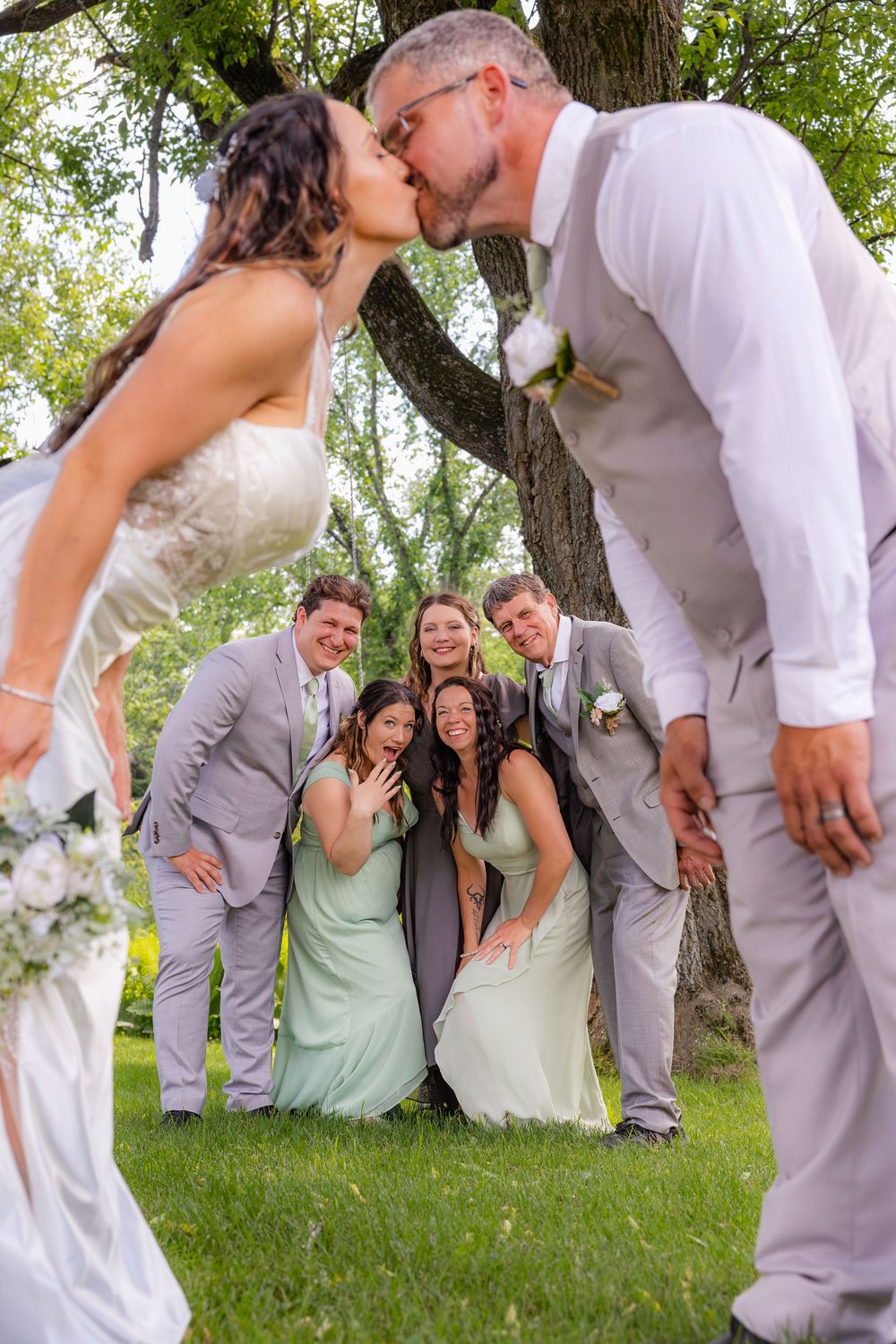 Wedding party in mint green and gray poses playfully behind a kissing couple in front of a tree outdoors.