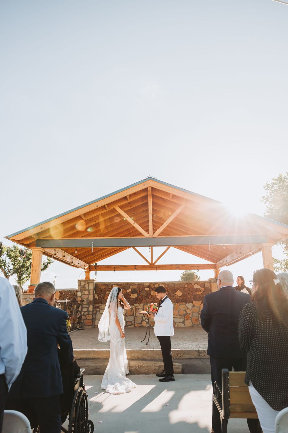 Golden sunset illuminates a wedding ceremony under a rustic wooden pavilion with stone walls and mountain views.