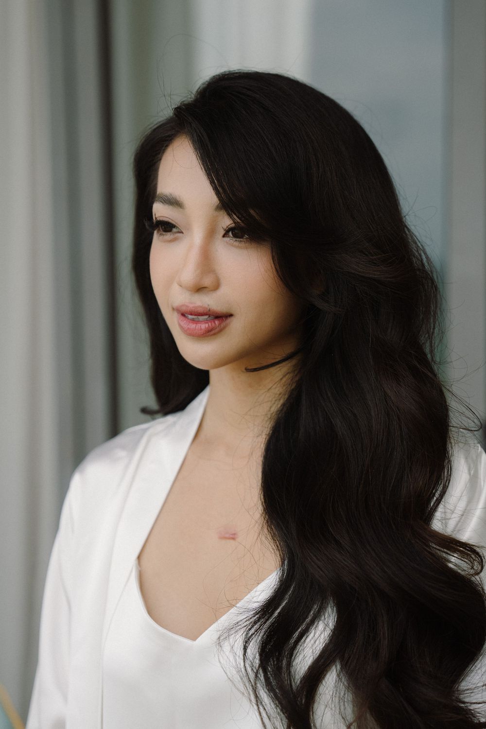 A woman with long dark hair poses in white clothing against a light background.
