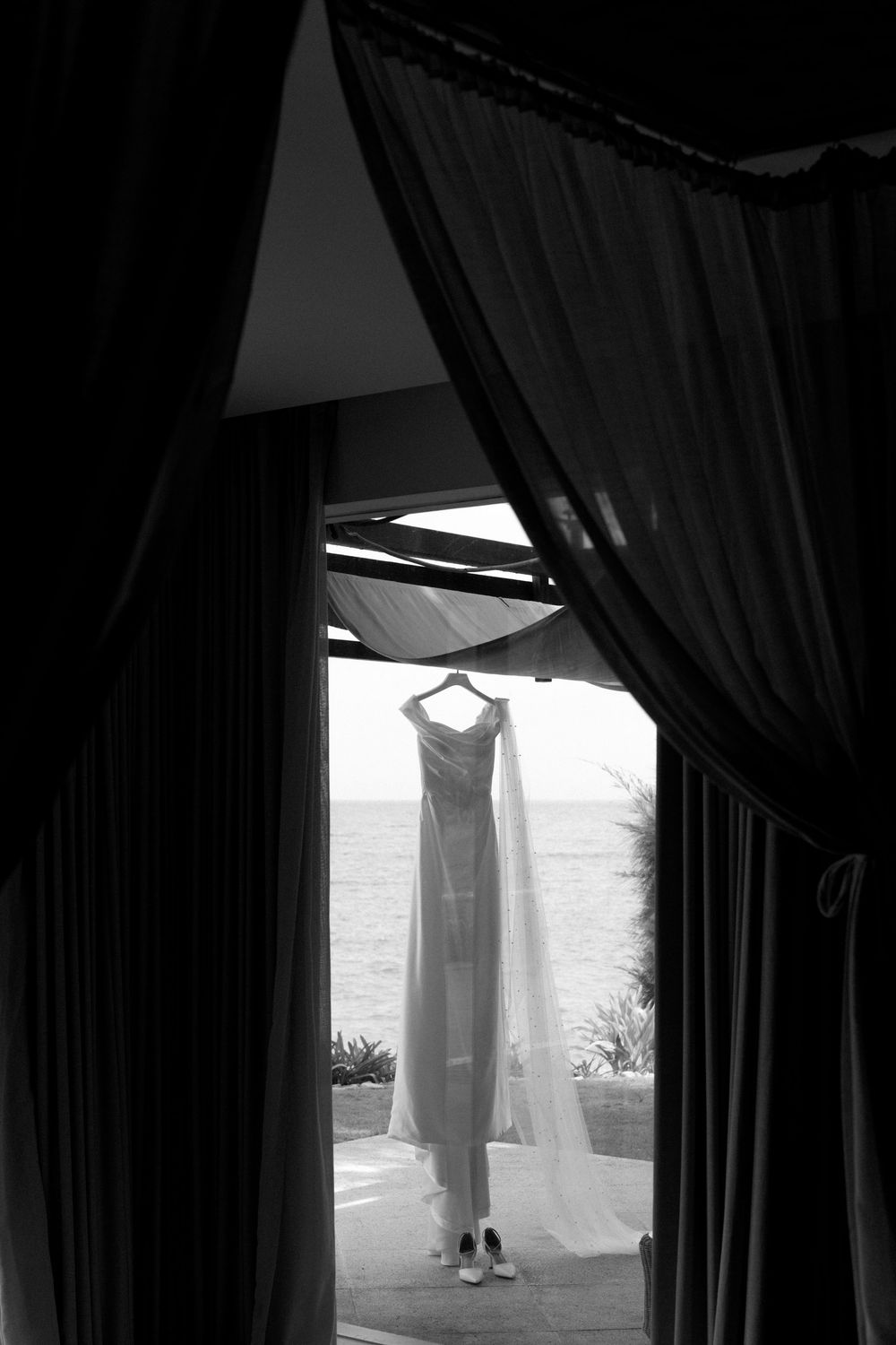 Silhouette of wedding dress hanging in window frame with ocean view.