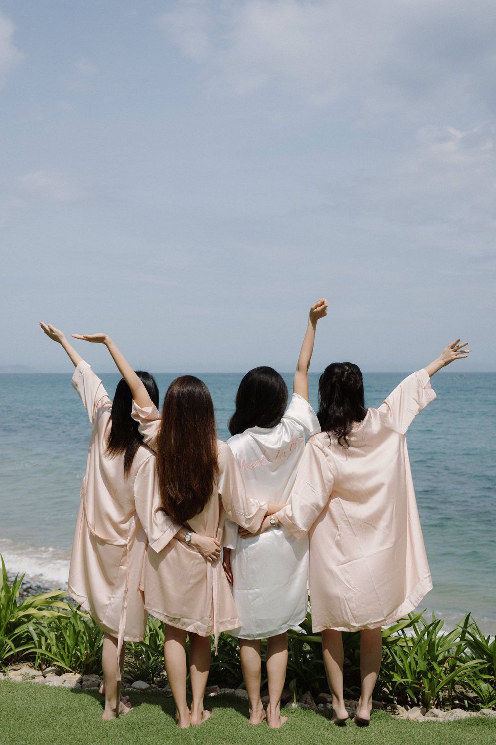 Four people in white robes stand with their arms raised overlooking a tropical ocean view.