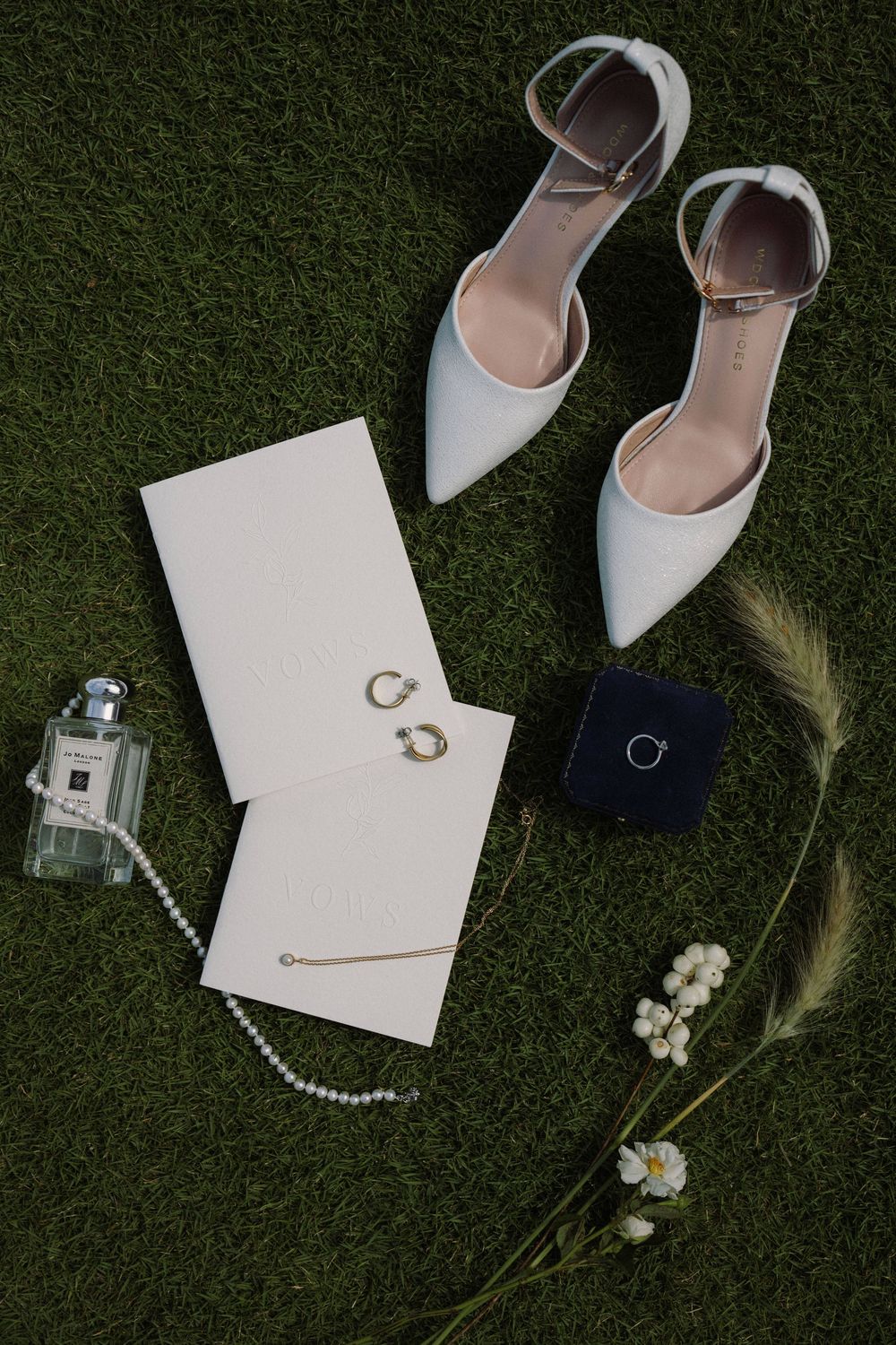 Wedding accessories including white heels, jewelry, and stationery arranged on dark grass.