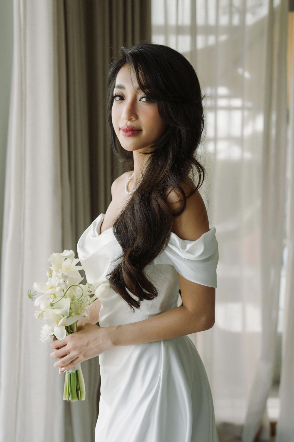 Elegant bride in white dress holding bouquet of white flowers in sunlit room.