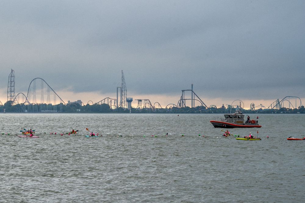 Small fishing boats and swimmers navigate misty waters with an industrial backdrop of Cedar Point amusement park
