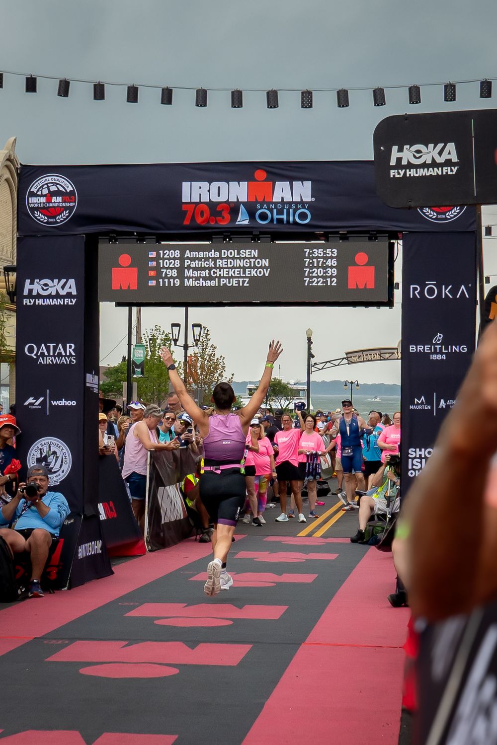 Multiple runners cross the finish line at an Ironman Ohio triathlon event with pink carpet and sponsor banners visible.