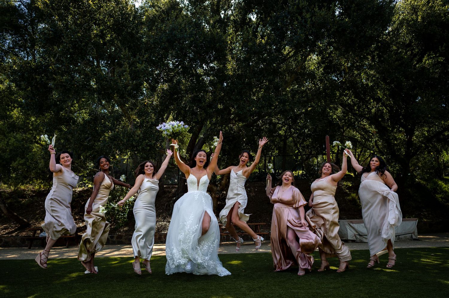 A group of wedding party members jump simultaneously in a synchronized celebratory pose on a sunny lawn.