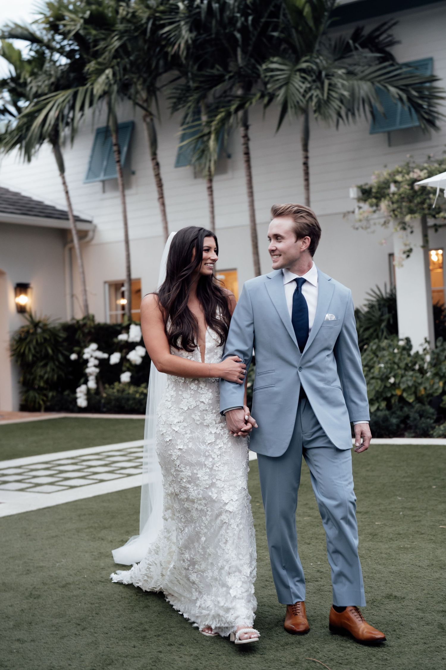 Newlyweds walk together on a lush lawn with palm trees and a white building in the background.