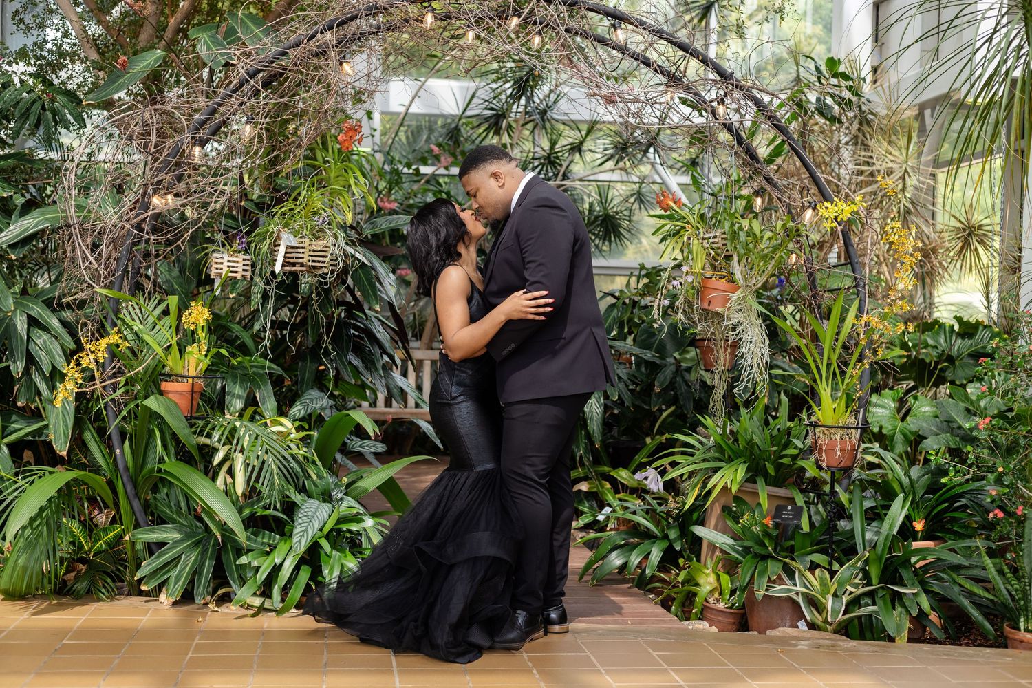 Romantic couple in formal attire embracing in greenhouse filled with tropical plants.