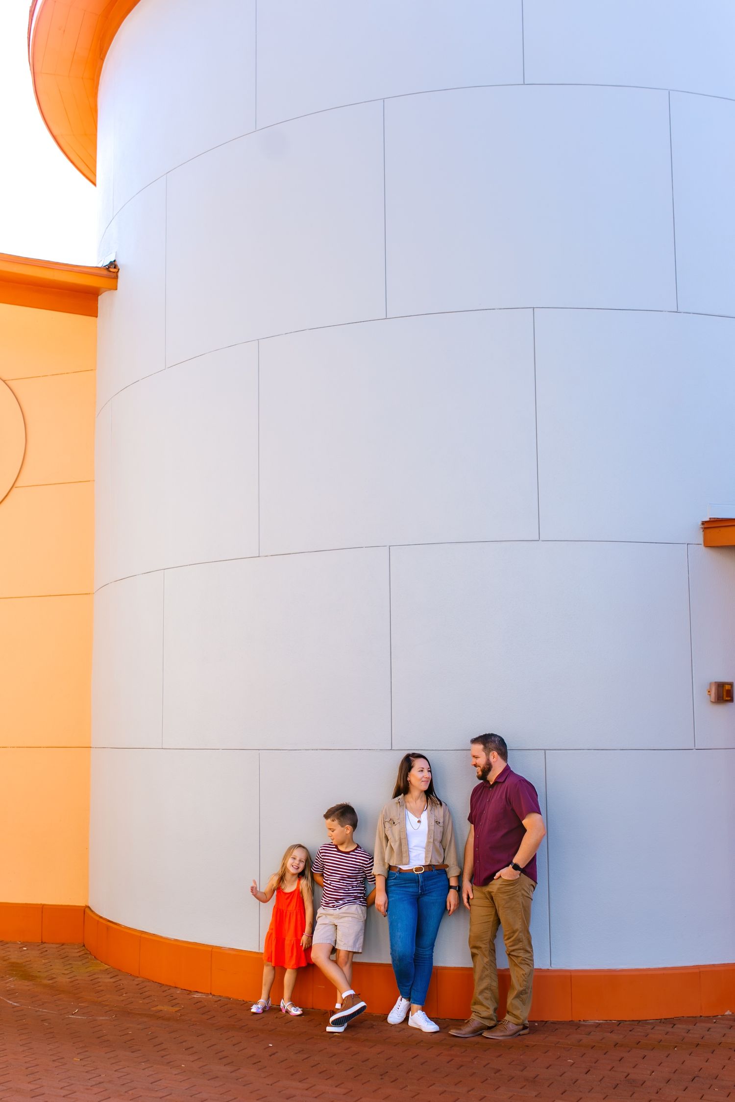 Group stands against large white cylindrical building with orange accents in bright sunlit setting.