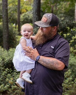 A bearded person in a cap holds a smiling baby wearing a white dress.