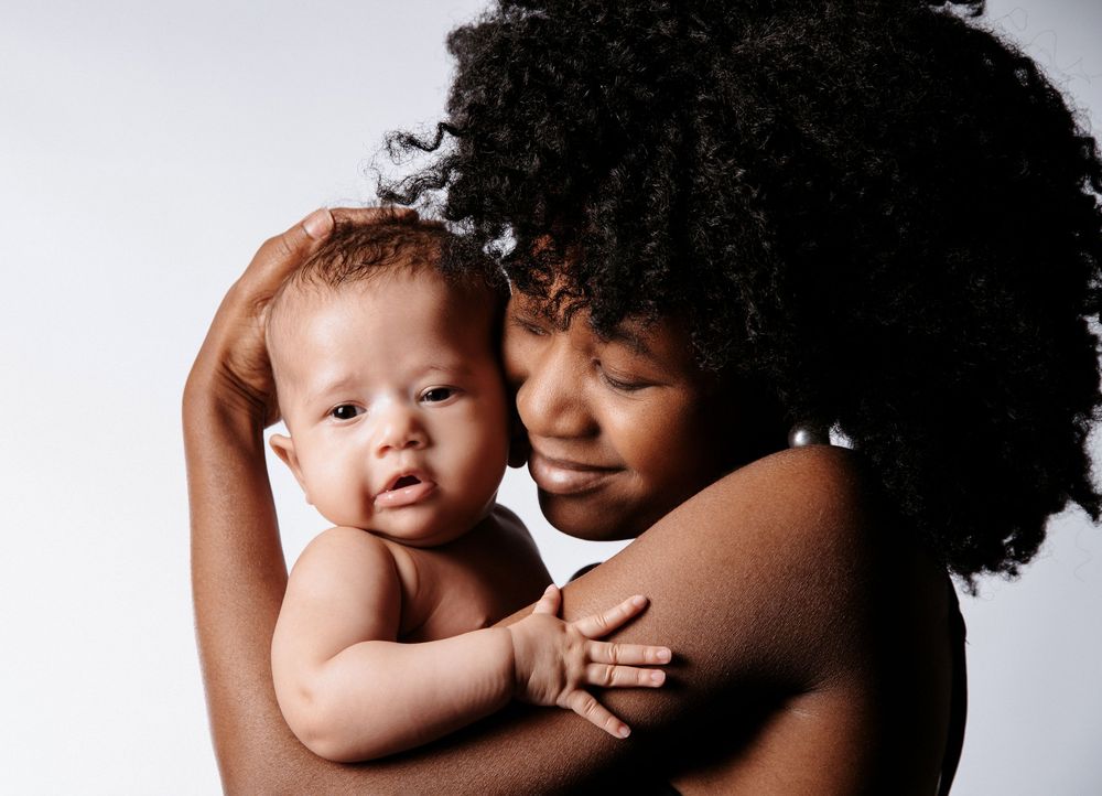 Farbfoto: Mama mit dunkler Haut hält ihr Baby, Hand schützend am Kopf. Liebevolle Babyfotografie in Gelsenkirchen voller Geborgenheit.