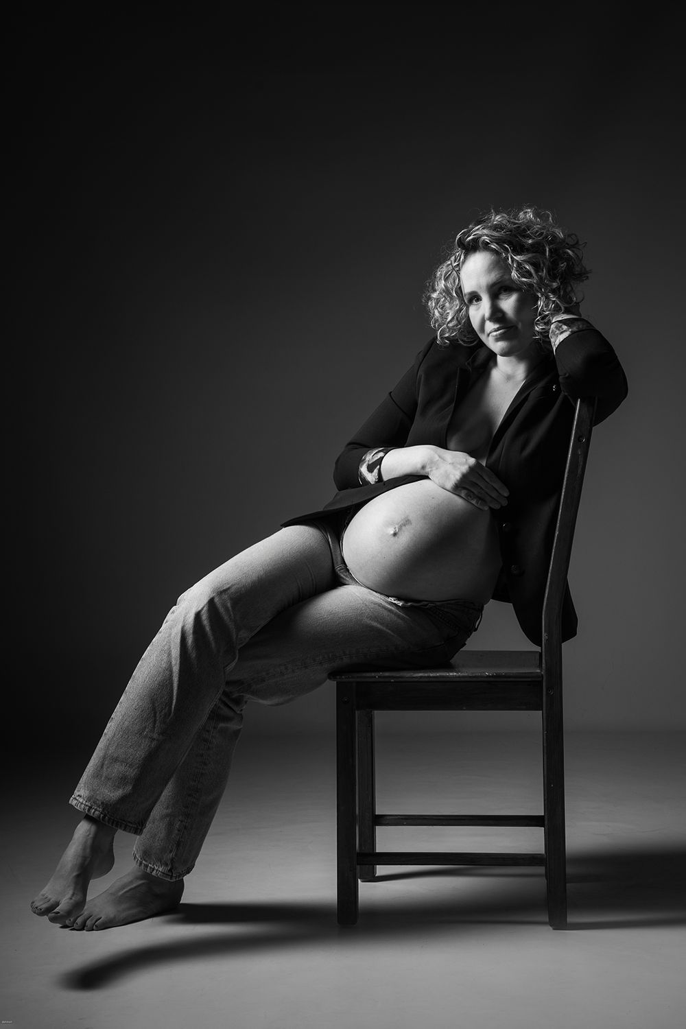 Schwangere im Anzugjacke und Jeans, Hand am Bauch, auf Stuhl in Schwarz-Weiß. Stilvolle Babybauchfotografie, Kokerei Hassel / Gelsenkirchen.