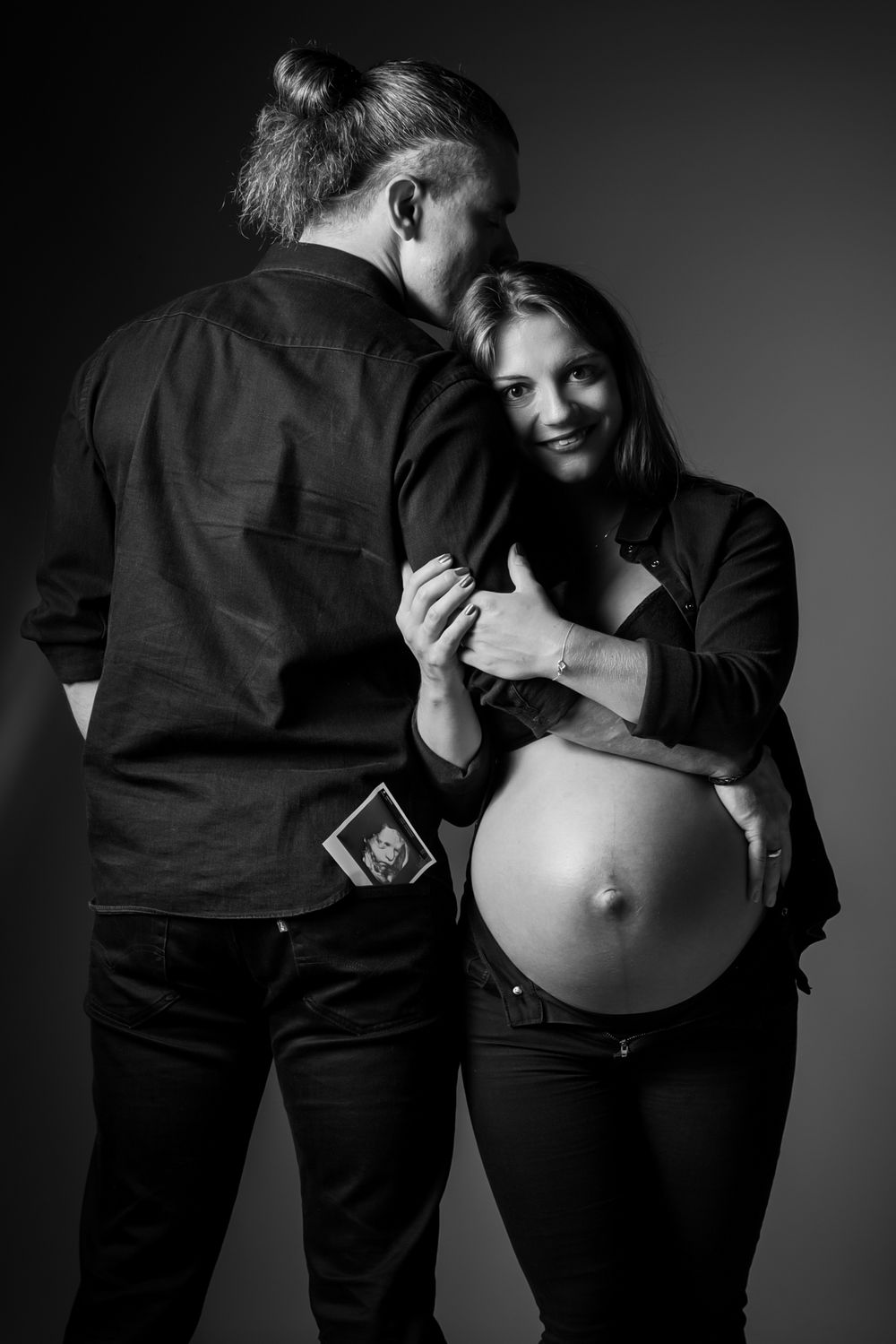 Ein Schwangerschaftsfoto in schwarz/weiß. Die Schwangere kuschelt sich an den Arm ihres Mannes. Ein glücklicher Moment werdender Eltern.