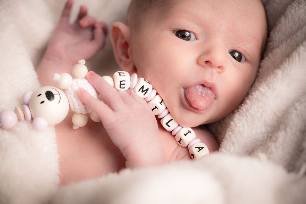Farbfoto: Baby liegt auf weichem Fell, hält eigene Schnullerkette, Zunge rausgestreckt. Kreative Babyfotografie im Fotostudio Gelsenkirchen.