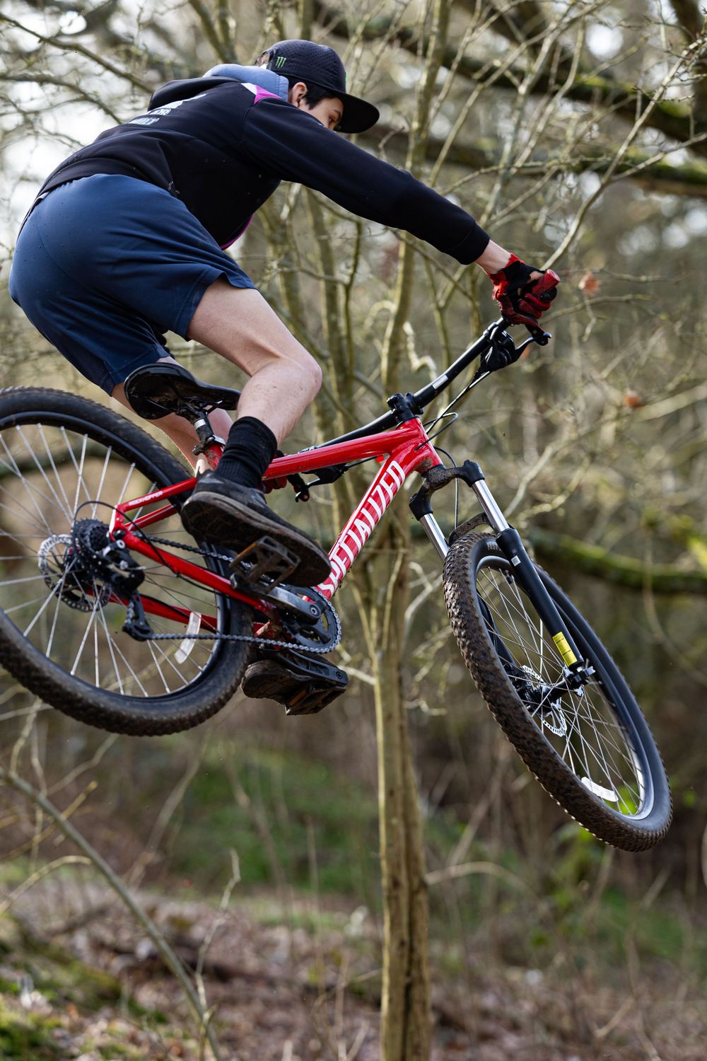 A mountain biker performs a jump sequence on a red specialized bike through a wooded trail area.