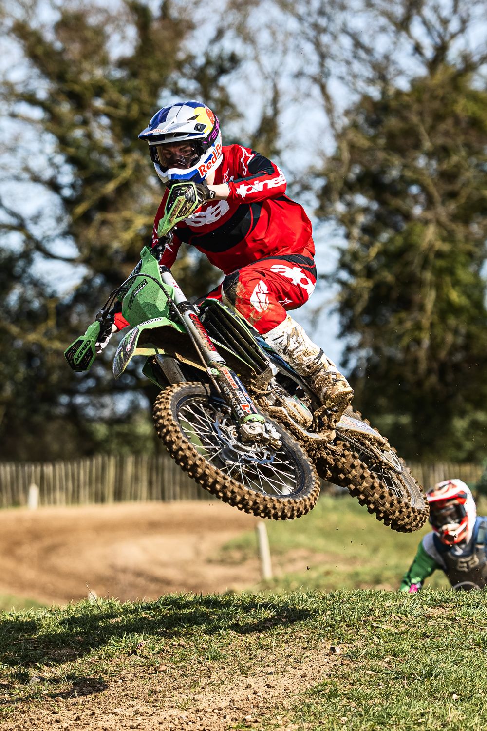 Motocross rider in red jersey performing a dramatic jump on a dirt bike over a dirt mound on a sunny day.