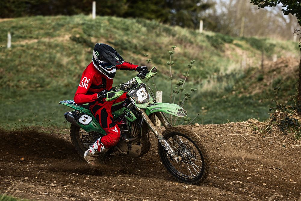 Dirt bike rider navigating a turn on a dusty motocross track with trees in the background.