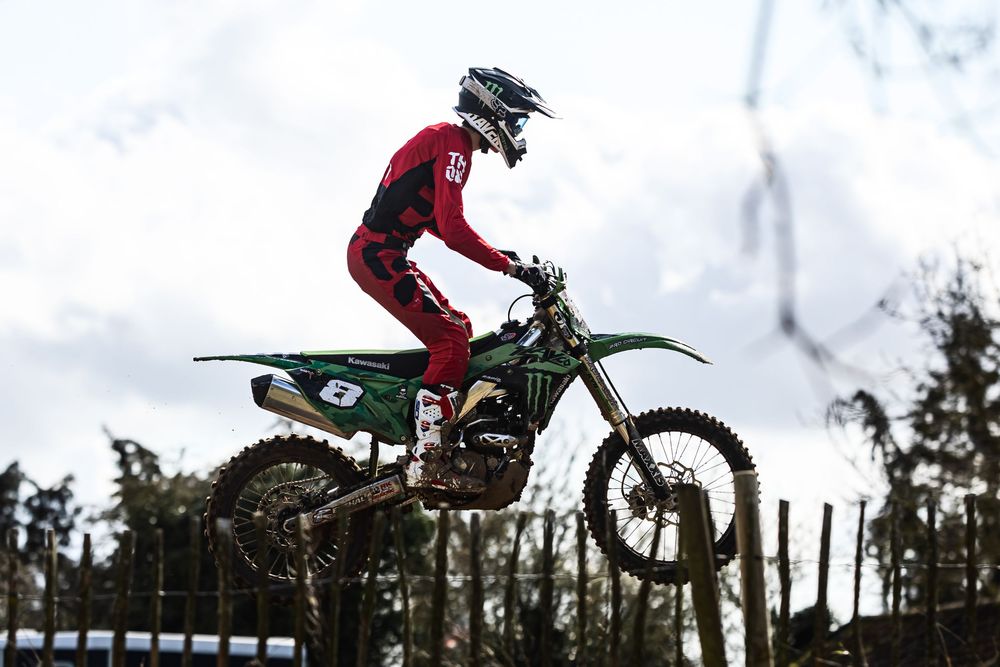 Motocross rider performing a jump sequence on a dirt track against a cloudy sky background.