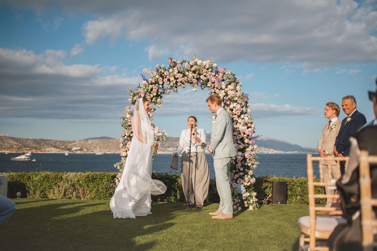 Outdoor wedding ceremony takes place under a floral arch along a stunning coastal backdrop as guests look on during golden hour.