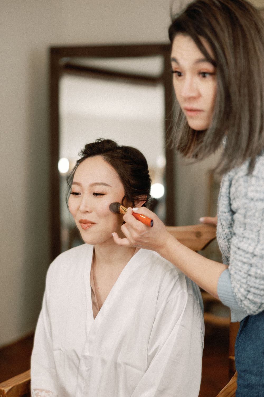Makeup artist applying blush to an Asian bride in a white robe before her wedding.