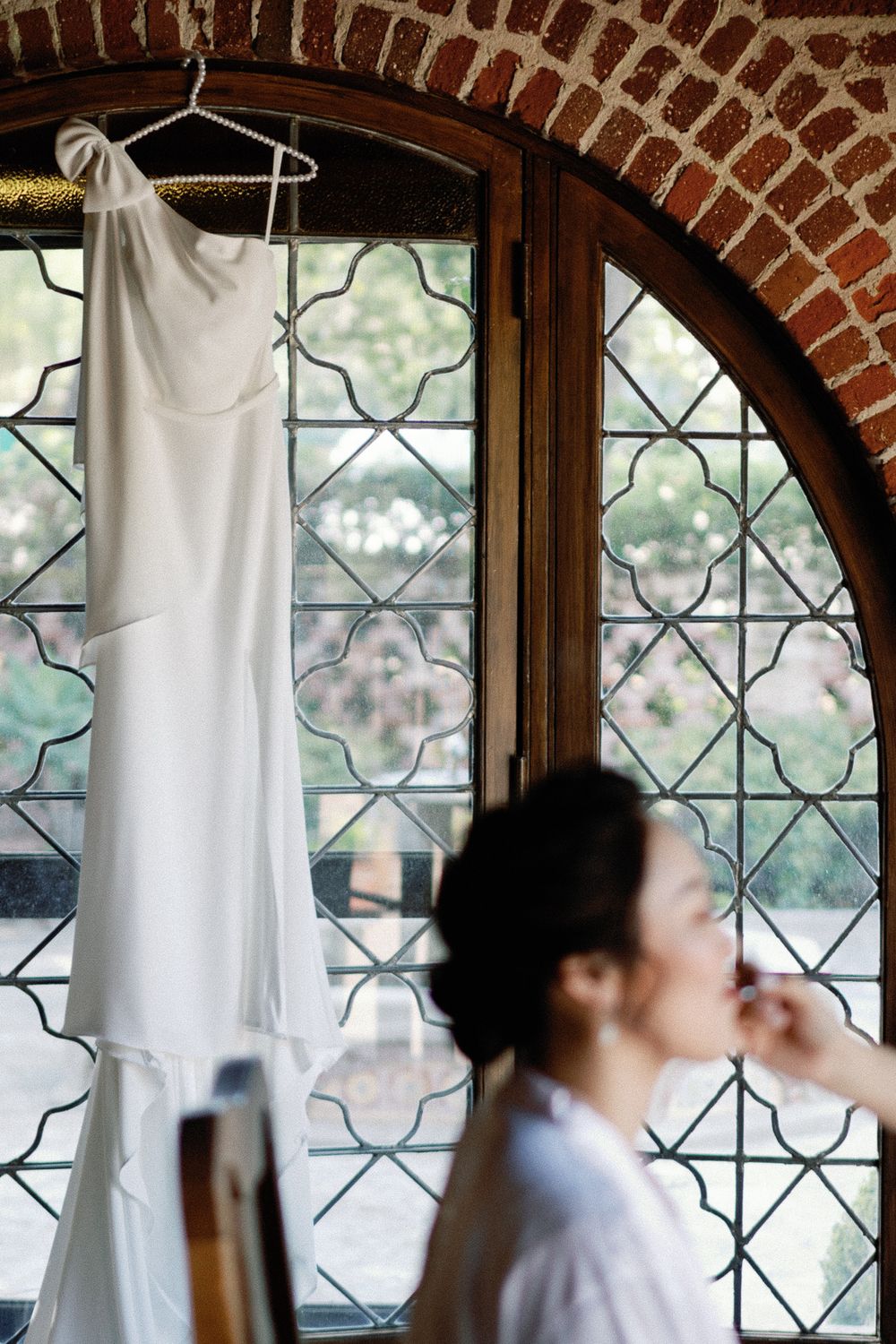 White wedding dress hanging in an arched stained glass window while bride gets ready below.
