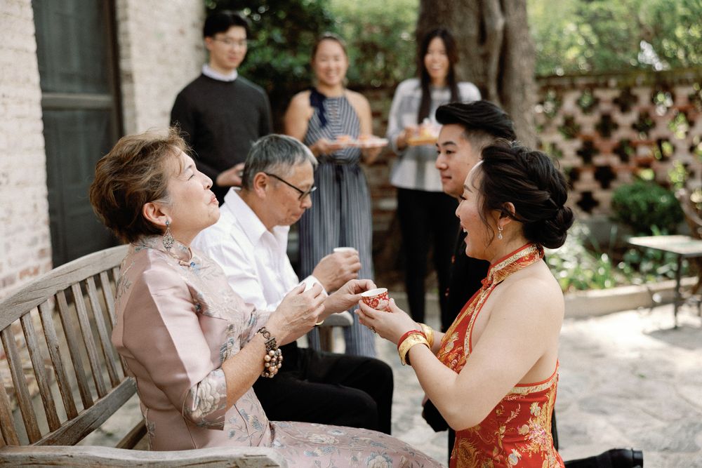 Bride in red qipao shares a joyful moment with seated guests during pre-wedding tea ceremony.