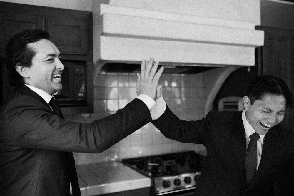 Two grooms in black suits laughing and giving a high five in a kitchen.