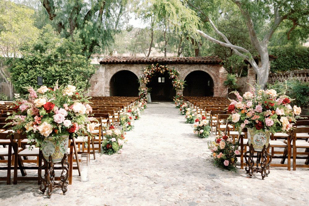 Elegant outdoor wedding ceremony aisle lined with lush floral arrangements leading to a rustic chapel.