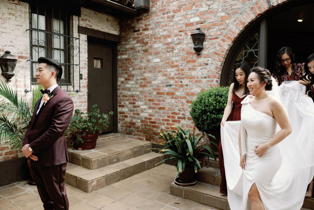 Bride in white gown and groom in burgundy suit share first look moment outside brick building.