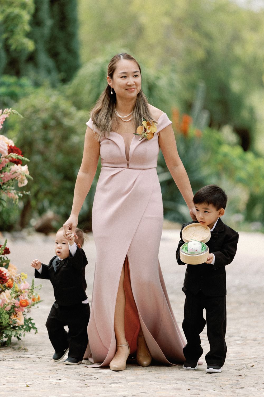 Bridesmaid in blush pink gown walks down aisle with two young ring bearers at outdoor wedding.