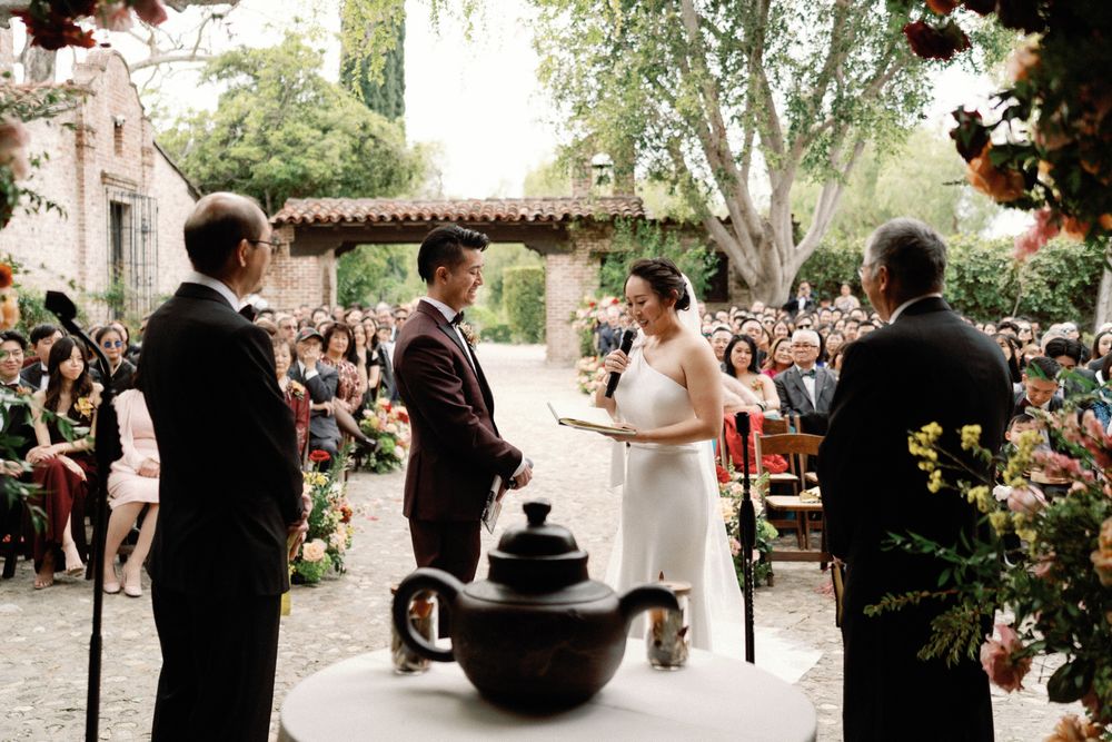 Couple exchanges vows at outdoor wedding ceremony with floral arch and seated guests.