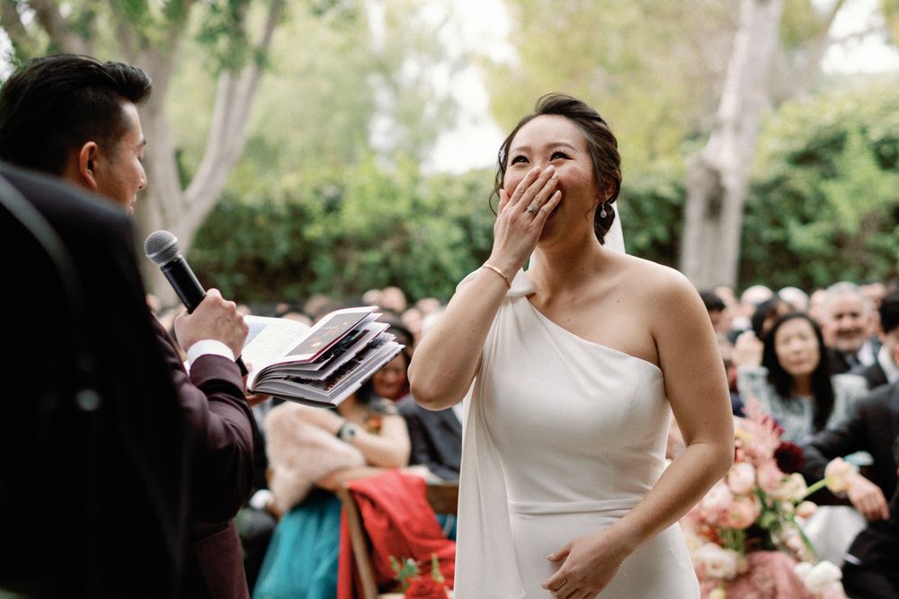 Emotional bride covers her mouth during outdoor wedding ceremony while groom reads vows.