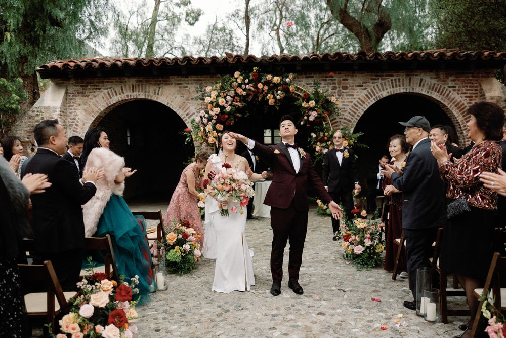Newlyweds celebrate joyful recessional through floral-lined aisle under brick archway.