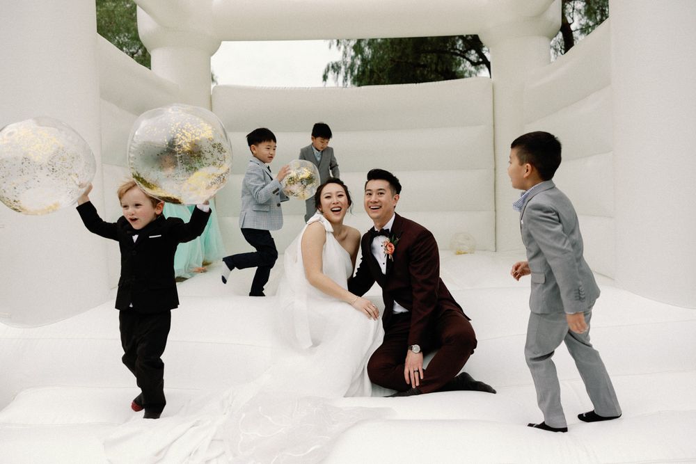 Bride and groom laugh with children on white bounce castle at outdoor wedding celebration.