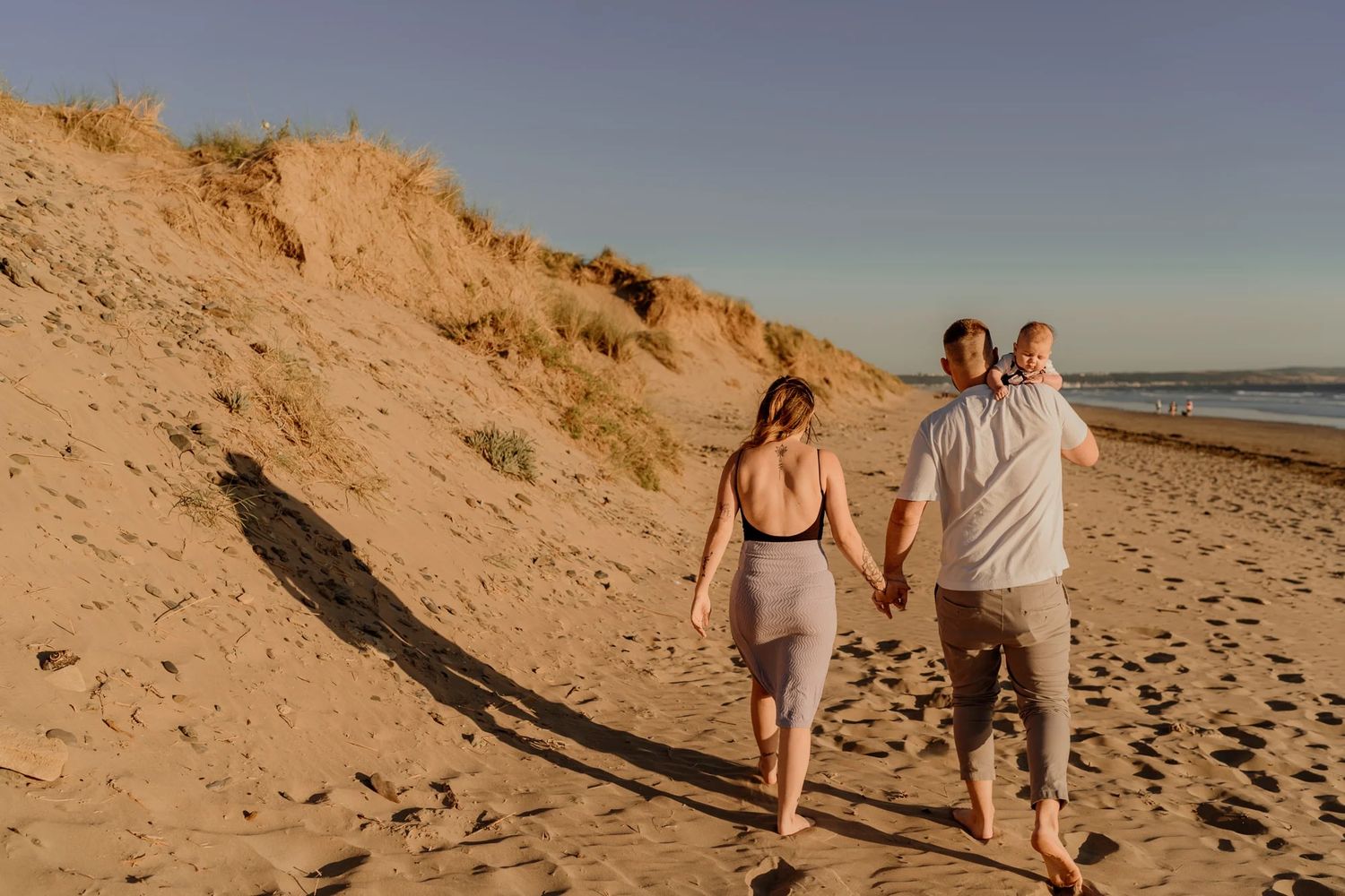 A couple walks hand in hand along a scenic beach with sandy dunes during golden hour sunset.