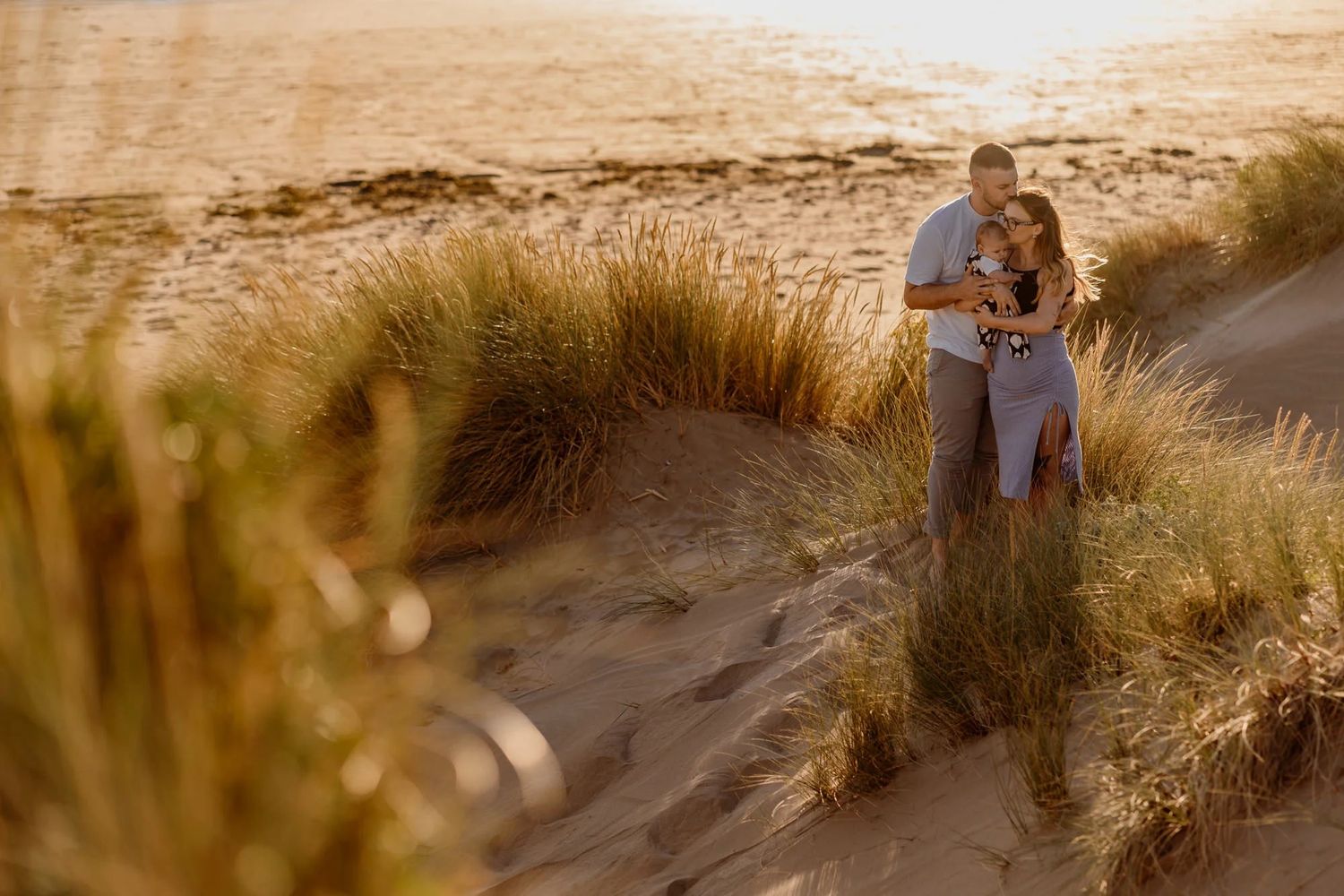Couple embracing among sand dunes and beach grass during golden hour at sunset.