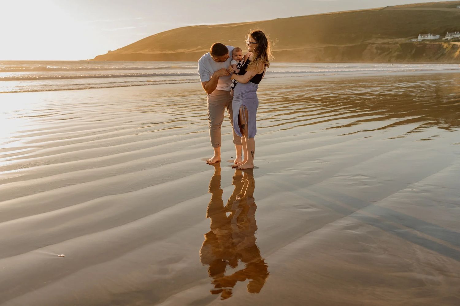 Family standing together on reflective wet beach sand at sunset with hills in background.