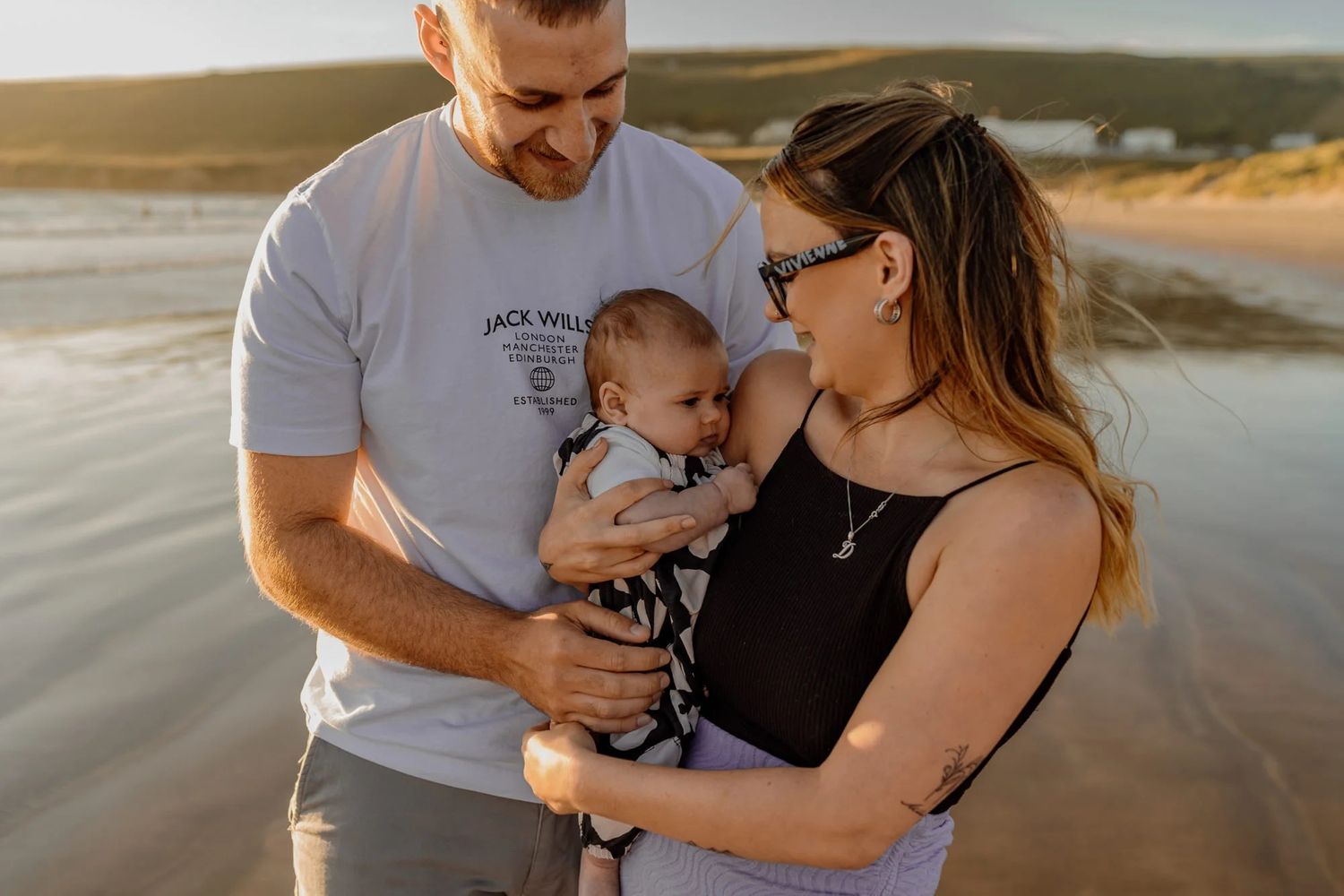 A couple holds their infant baby together while standing on a beach at sunset.