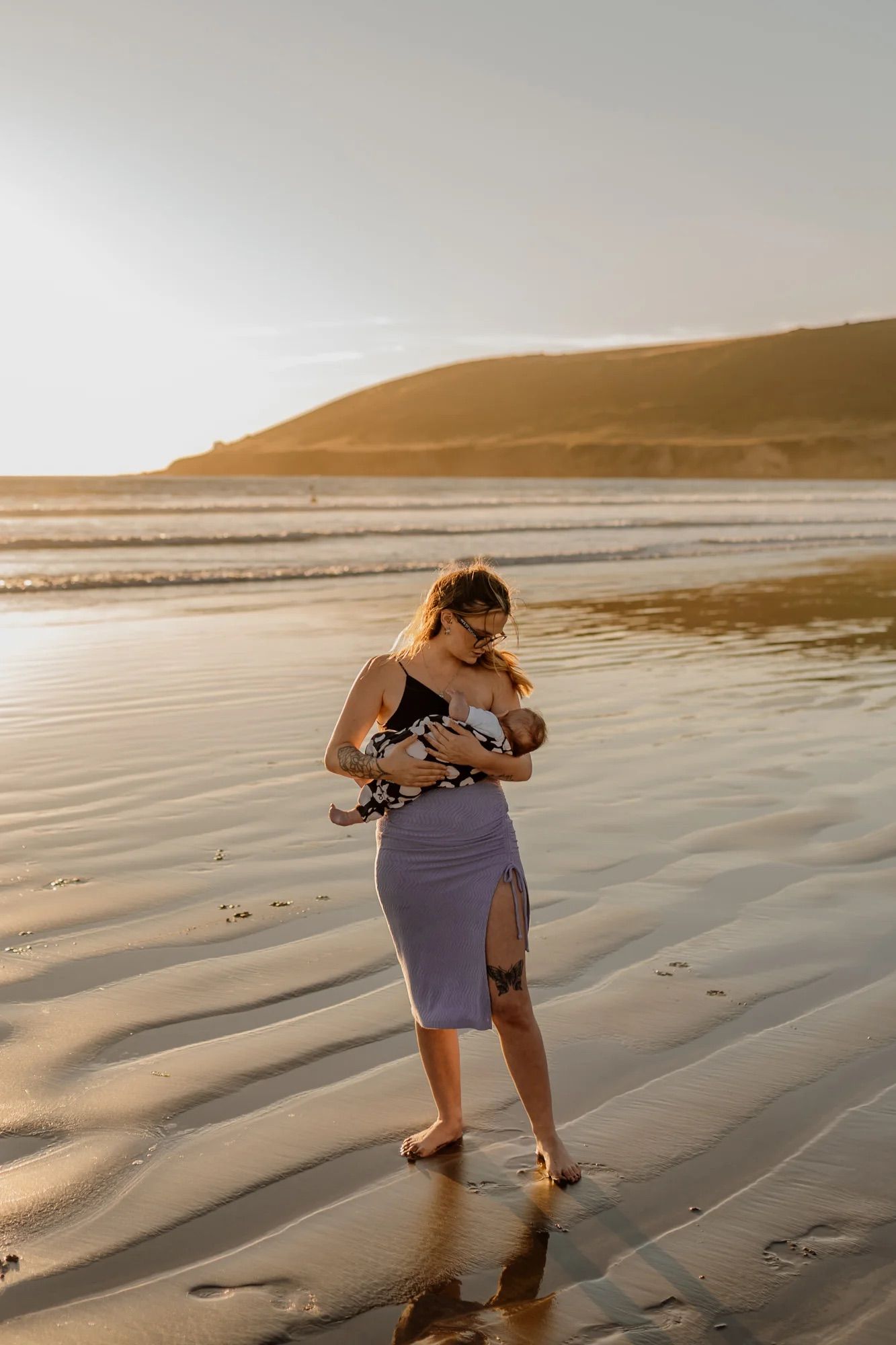 Figure in a purple skirt stands alone on a beach at golden hour holding something close to their chest.