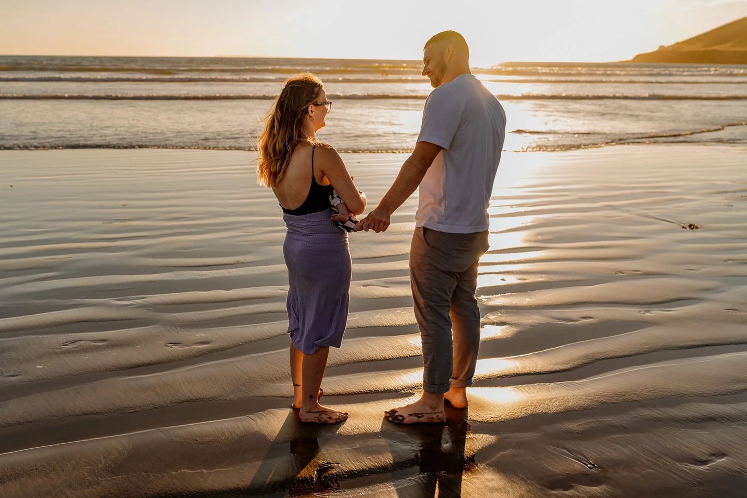 Two people stand together on wet sand during sunset at the beach.
