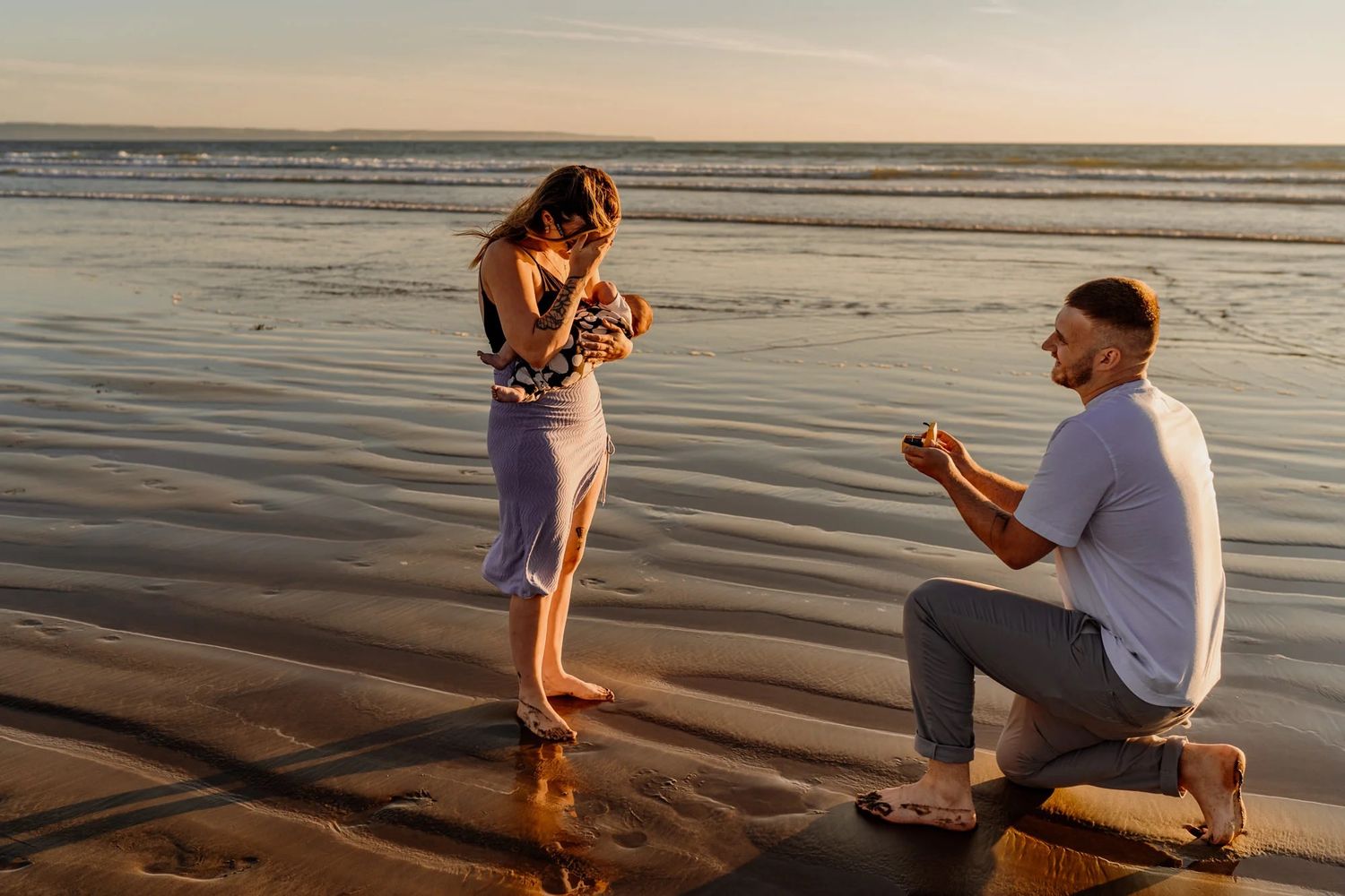 A surprise proposal moment unfolds on a scenic beach at sunset with waves in the background.