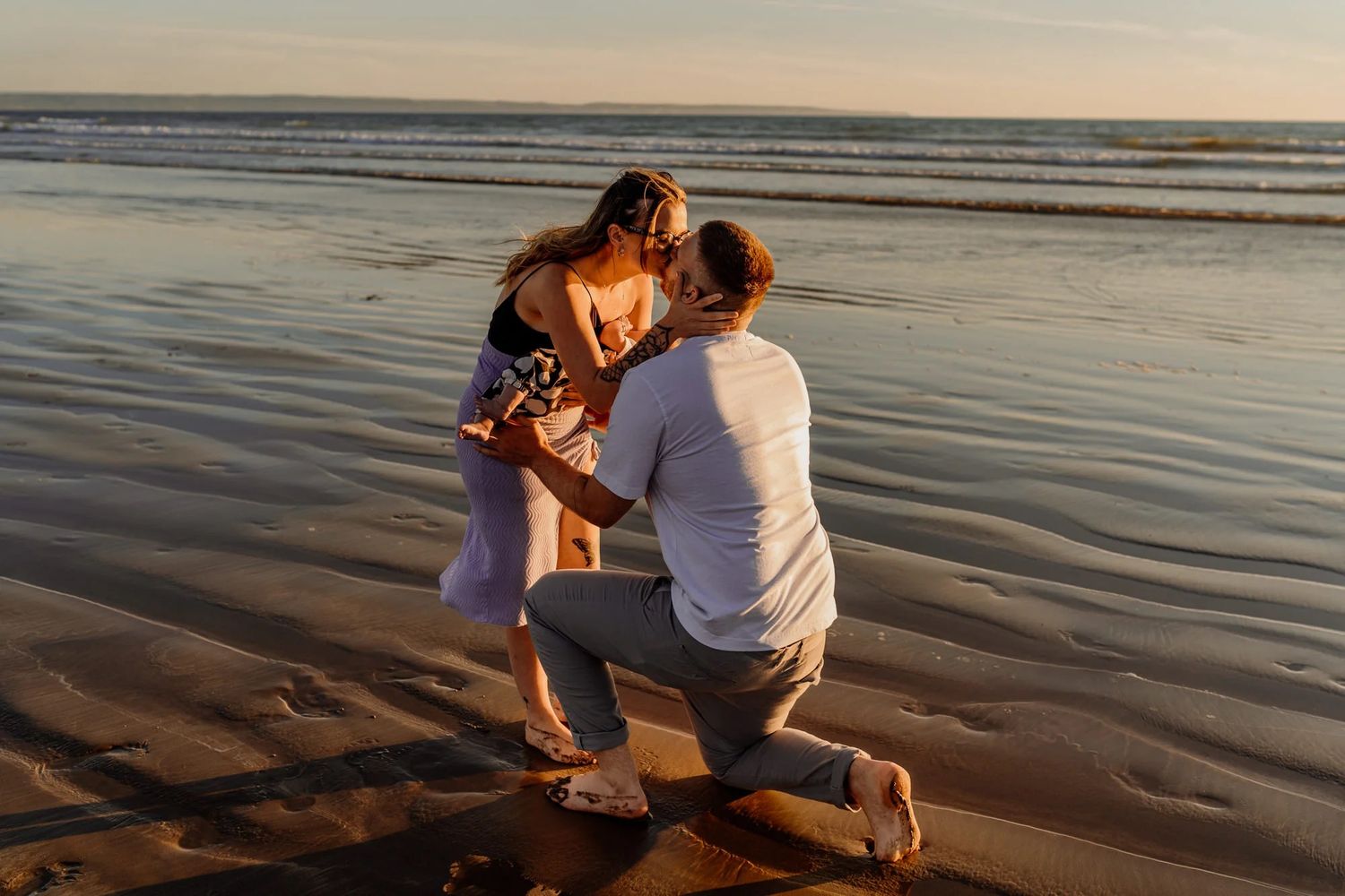 Couple embraces on sandy beach at sunset with soft golden light reflecting on calm ocean waters.