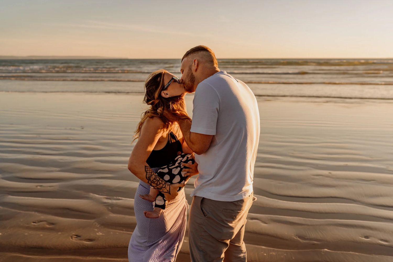Couple embraces tenderly on a serene beach at sunset with gentle waves and golden light.