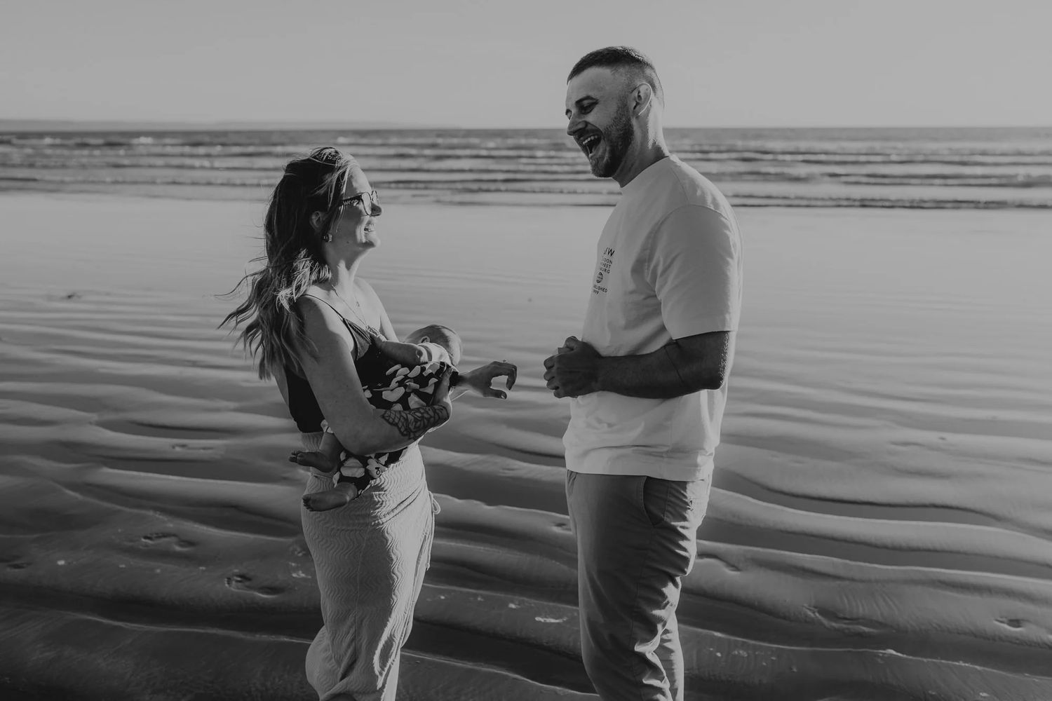 Black and white photo of couple sharing an intimate moment on beach at water's edge.