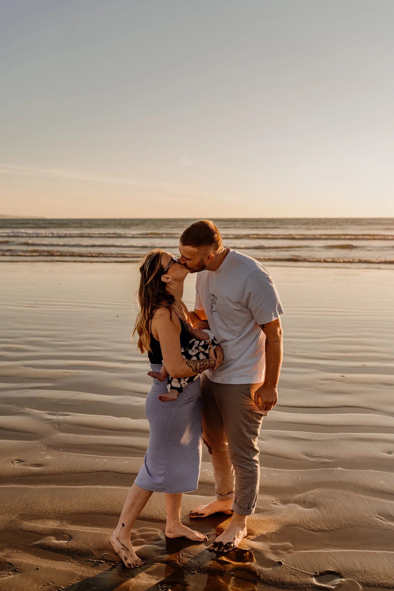 A romantic couple embraces on a scenic beach at sunset with gentle waves and sandy shore in the background.